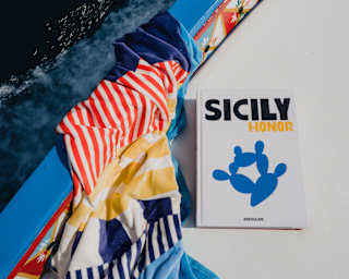 A copy of Sicily Honour rests on the deck of a boat with a pretty, hand-painted side, next to a red, blue and yellow towel.