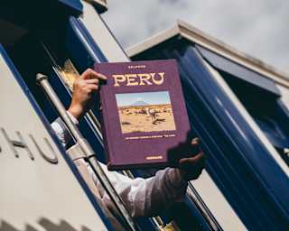 Low-angle image looking up at a train carriage doorway where a man's arms hold out a copy of the Belmond publication, Peru.