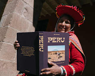 A Peruvian woman in brightly-woven traditional clothing holds open Belmond's travel book, Peru, with an alpaca cover-image.