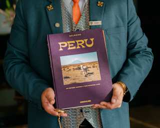 A man in a teal suit with gold hints and a patterned waistcoat holds a front-facing copy of Peru, by Belmond and Assouline.