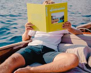 A man reads Splendido by Belmond and Assouline, obscuring his face with the open book as he relaxes aboard a boat at sea.