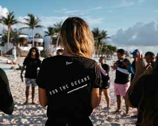 A ring of people wearing Parley For The Oceans tops gather on the beach, seen close-up from behind a blonde woman.