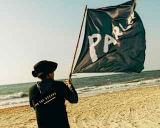 A man in dark clothing and hat stands on a beach with a large black flag with the word PARLEY in white, seen from behind.