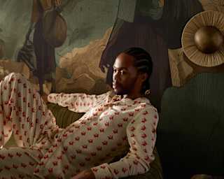 Thebe Magugu reclines in a hotel suite he designed, seen in low light wearing his Men’s Vanilla and Red Monogram Set.