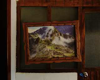 A photograph of Machu Picchu covered with floss-like clouds hangs lopsided in a wooden frame on a wall with glass panels.