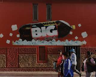 People walk past a building painted red with a mural of a bottle on its side on a layer of ice advertising Mega Big Cola.