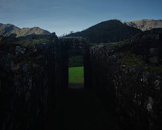 The doorway in the shadowy Llactapata stone ruins reveals a verdant grass area with a backdrop of mountainous ridges.