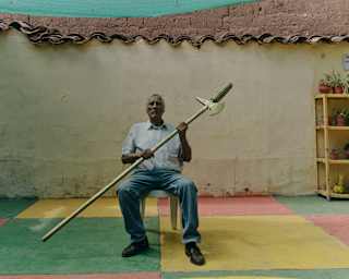 Alfredo Inca Roca, descendant of the Inca Viracocha, holds a golden spear in shirt and jeans, seated in a rustic courtyard.