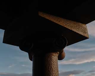 An ancient column's capital and architrave stands tall against a moody cloud-strewn sky of blue and grey, in close-up.