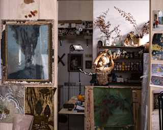 Elena Carozzi, with chignon-ed blond hair looks for pigments on an art shelf, seen from behind in her busy wallpaper studio.
