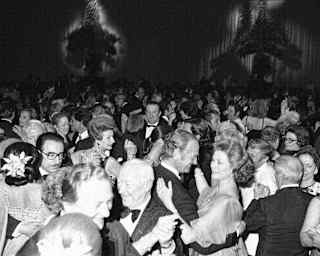 A black and white photo shows a large crowd of people dressed formally, dancing closely together at a party or event. The background features dim lighting and decorative wall shadows.