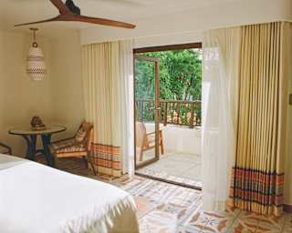 A Deluxe Garden View Room, seen from the bedside, with table and cane chairs, traditional tiling and open doors to a balcony.