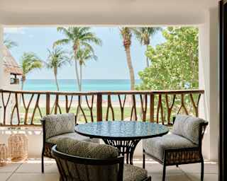 A table and three upholstered chairs sit on a Master Suite balcony with carved wood railing, claiming sublime beach views.