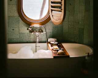 A shutter opens to reveal sea views through an oval window set in the wall above a bubbling bath with a cross-tub soap tray.