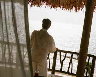 High angle view of a man in white top and trousers leaning on his balcony rail as he gazes at sun-bleached sea below.