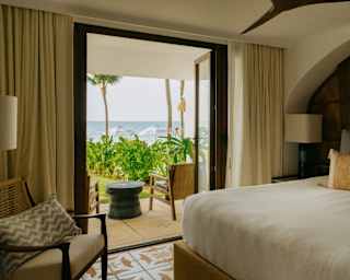 Open bedroom doors reveal a terrace, garden and immediate ocean views, seen over a large plump white bed in Villa Mariposa.