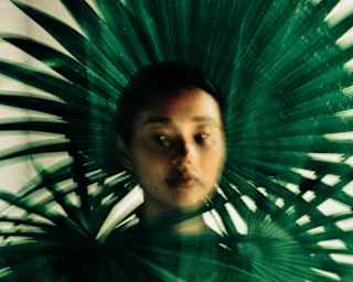 Artistic long-exposure image of a woman's face surrounded by fan palm leaves, with one like a flamboyant headdress behind.