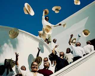 Smiling male and female members of Maroma's staff crowd onto a white stairwell, tossing Panama hats into a deep blue sky.