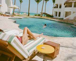 A lone swimmer enjoys the rippling blue tropical pool, seen over a lounger where a female sun-bather's legs stretch out.