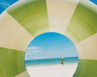 A man runs across a white beach by an azure sea, holding a yellow kite in his hand, viewed through an inflatable rubber ring.