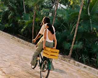 A woman rides on the back of a bike, holding onto a yellow box behind her as the male rider sets off along the stone path.