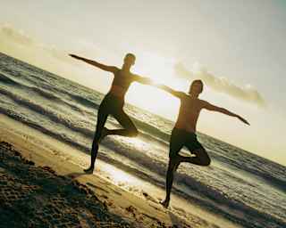 Angled image of two men in wide-armed tree poses, half-silhouetted against the sea and sun, as they practice beach yoga.
