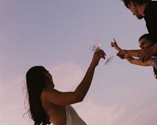 Angled shot of a long-haired woman in a white dress as she raises a glass with two male friends, taken against a lilac sky.