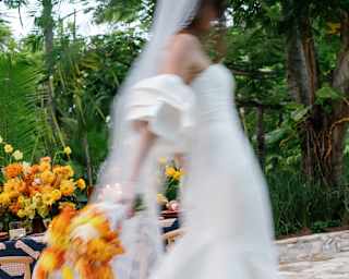 A bride in a long dress and veil creates a beautiful wedding-white blur as she walks with a bouquet of bright orange flowers.