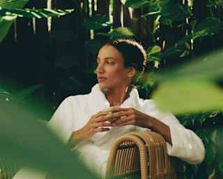 A woman in a white robe relaxes on a cane chair with a drink in the Maroma Spa by Guerlain courtyard, viewed through foliage.
