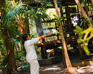 A person dressed in white stands outdoors among lush green trees, holding and wafting a smoking object, possibly performing a ritual or ceremony near a wooden structure. Sunlight filters through the foliage.
