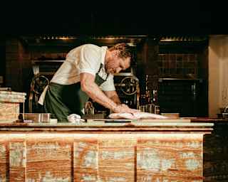 Chef Curtis Stone, in a white top and green apron, prepares a pink snapper on the countertop of a marble block at Woodend.