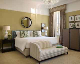 A pale chaise-longue abuts a crisp white bed with floral cushions and mirror above, in a mellow Deluxe Room with olive walls.