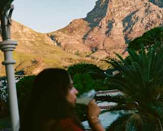 A dark-haired woman in red drinks a cocktail in soft-focus foreground, gazing at a sunny mountain backdrop from her balcony.