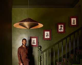 A person in brown clothing stands by a green staircase under a pendant lamp, with framed art on the wall and patterned carpet below.