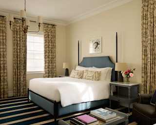 A large bed with blue valance and headboard rests in the centre of an airy Garden Cottage bedroom with three windows.