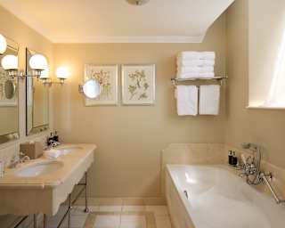 Three globe chrome lights divide two bevelled mirrors above a marble double sink unit in a cream bathroom with a white bath.