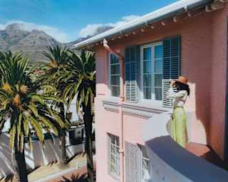 A woman in a brown hat, white shirt and green trousers stands on her suite balcony looking at the view through binoculars.