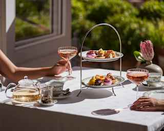 Two guests, out of shot, enjoy glasses of rosé at an Afternoon Tea table with a two-tier patisserie stand, in bright sunshine.