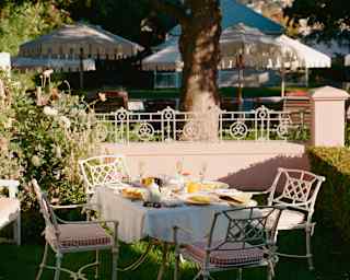 A breakfast table with four chairs, busy with plates and glasses, sits on the lawn in a cottage garden awaiting guests.