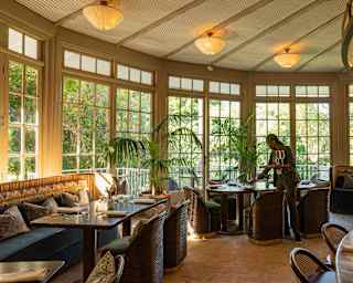 A waiter sets glasses on a table, preparing to welcome guests to Amura's garden-room style space with warm lights and plants.