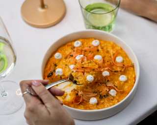 A guest lifts a spoon of an unctuous risotto-like dish dotted with cream, seen close-up from above at a table with water.