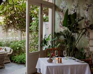 A table for two sits in a corner of The Verandah against green botanic-print wallpaper and potted plants, by a terrace door.