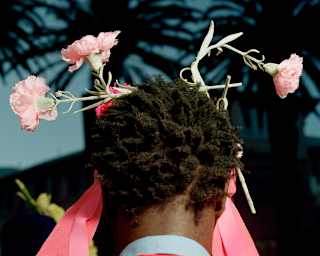 Pink carnations and ribbons adorn a model's head as he wears a red jacket and blue shirt by ChuSuwannapha, seen from behind.
