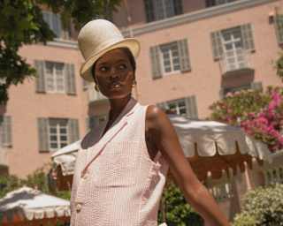 A female model poses in gardens in a Wanda Lephoto soft-stripe pink sleeveless top and trousers with a tall cream bowler hat.