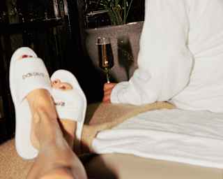 The slippered feet of a woman lying down rest on the end of a bed where a friend sits in a robe, with wine, seen from behind.