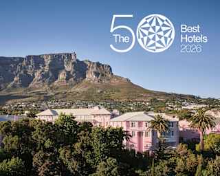 A pink hotel surrounded by trees stands before a large rocky mountain under a clear blue sky, with The 50 Best Hotels 2026 text in the upper right corner.