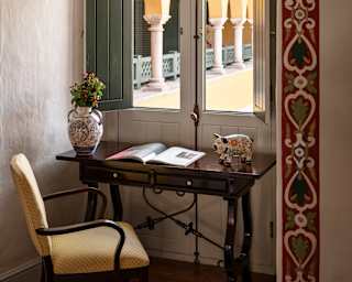 A desk and chair sit in an alcove with painted borders up against closed rustic doors with window panels and green shutters.