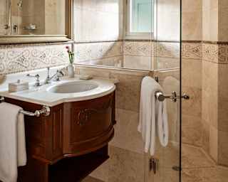 View of a Junior Suite bathroom in cream marble with a glass-fronted shower cubicle behind a white basin in a wood unit.