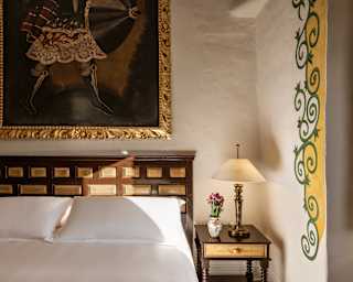 In a corner of a Junior Suite, sunlight plays across a white bed, illuminating gilt panels in the headboard and side table.