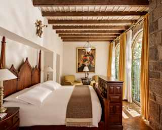 Sun spills through two arched windows into a bedroom with stone and painted walls, viewed over a bed below a beamed ceiling.